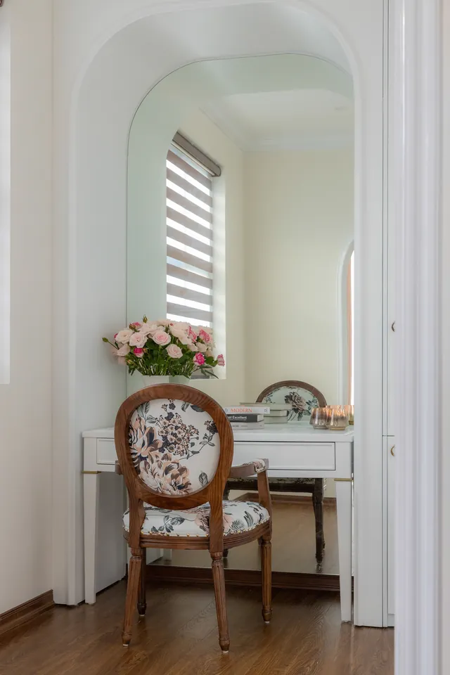 White dressing table with one drawer set into an arched mirrored alcove, with a floral upholstered oval-back chair and a vase of pink roses