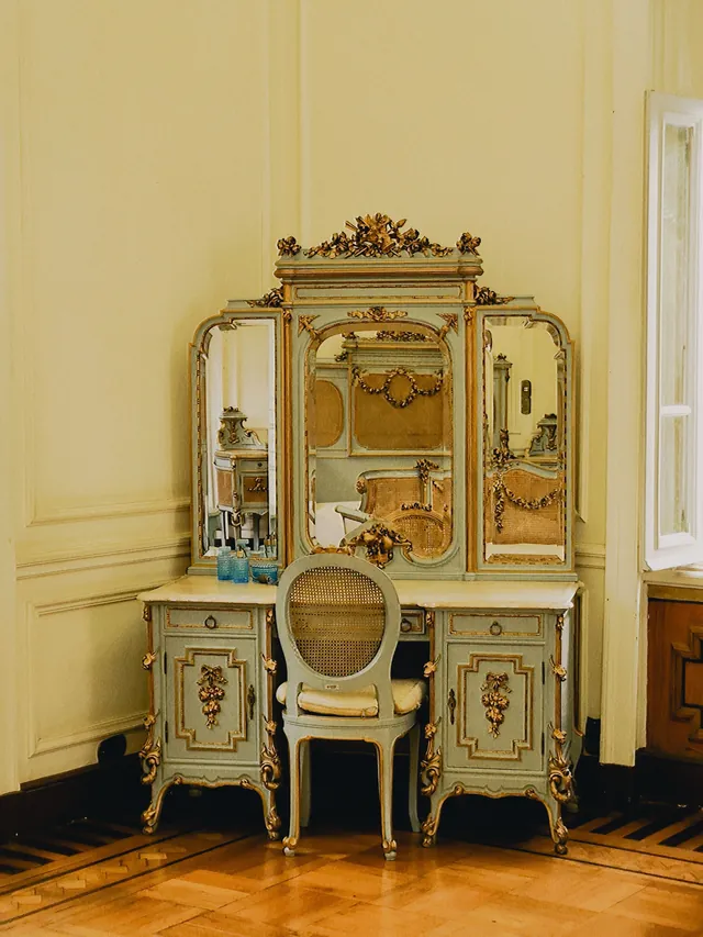 French-style painted vanity with a carved, gilded triple mirror, twin cabinets with doors and curved legs, with a matching cane-back chair
