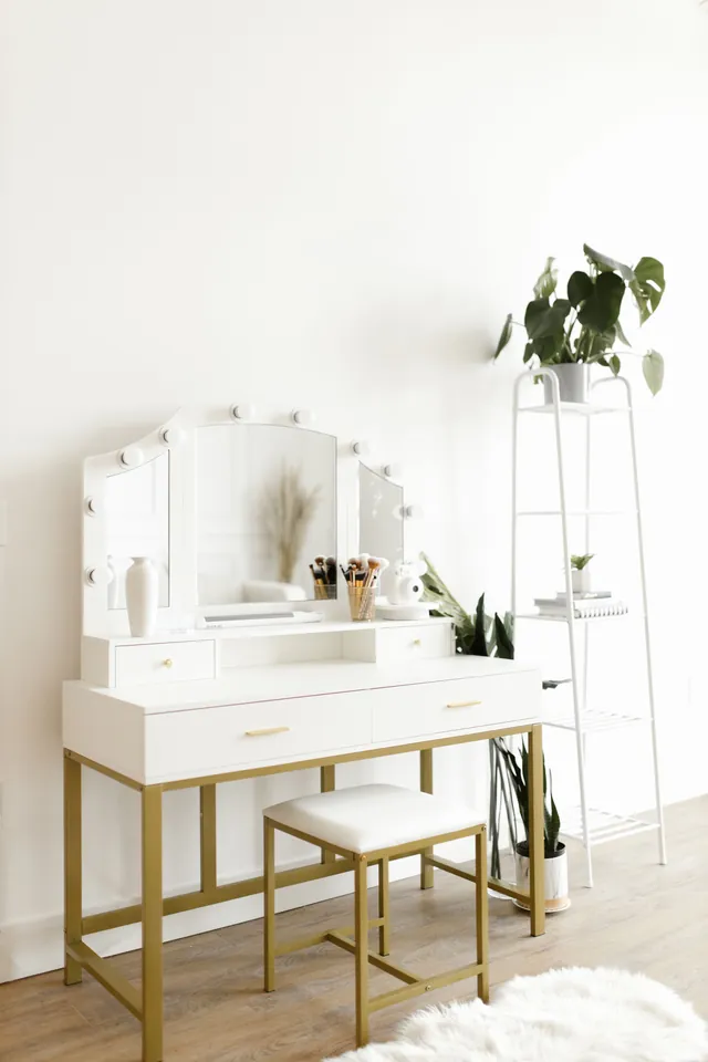 White vanity table with a lighted tri-fold mirror, small upper drawers, two main drawers with gold-tone handles and a matching upholstered stool