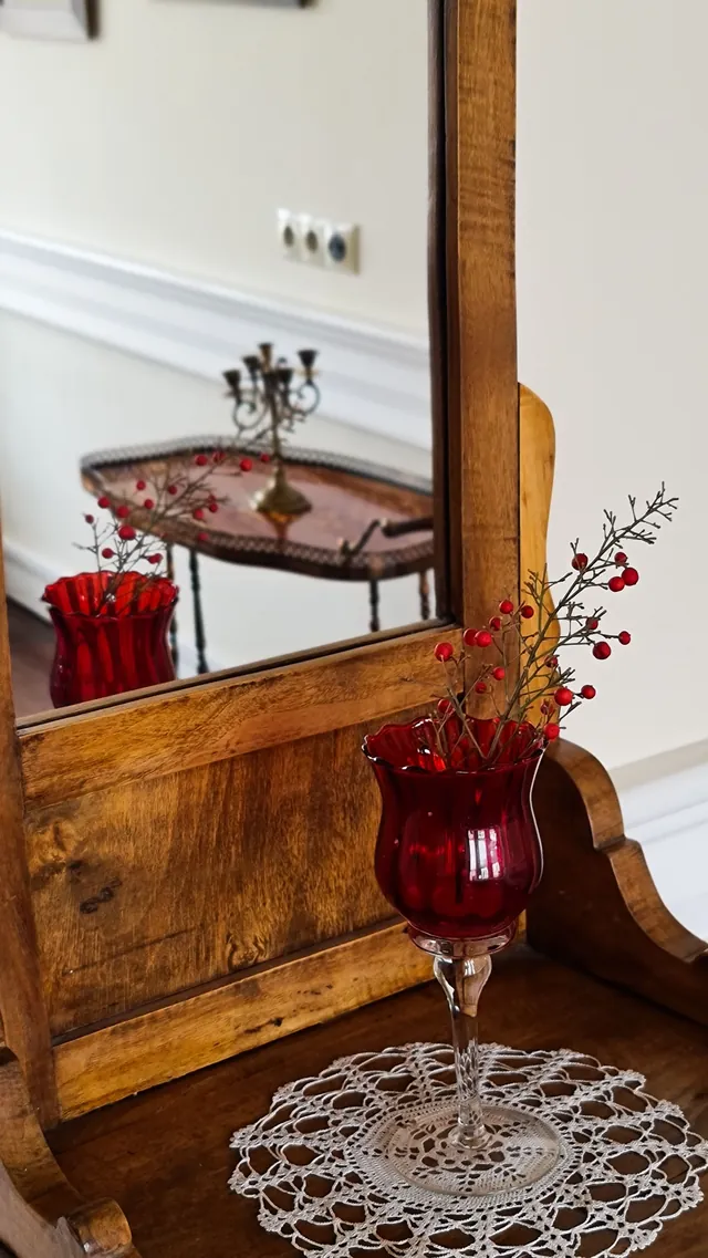 Close-up of a wood-framed dressing mirror with a red glass candle holder and berry sprig on a lace doily
